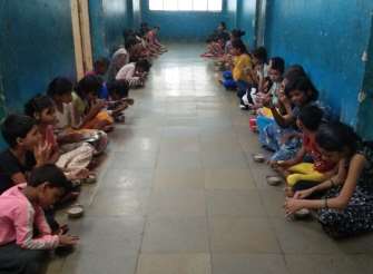 A photograph of two dozen girls sitting along the walls of a corridor, each with full bowls before them.